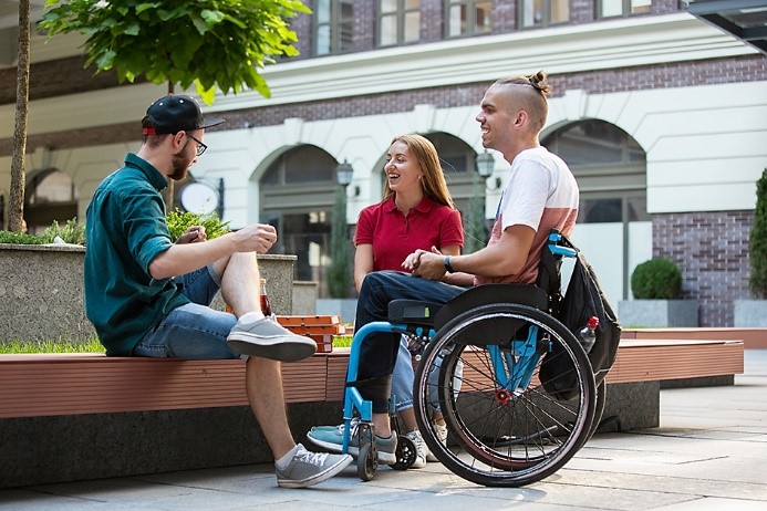 A young person seated in a wheelchair and two young persons seated on a bench cheerfully chatting altogether